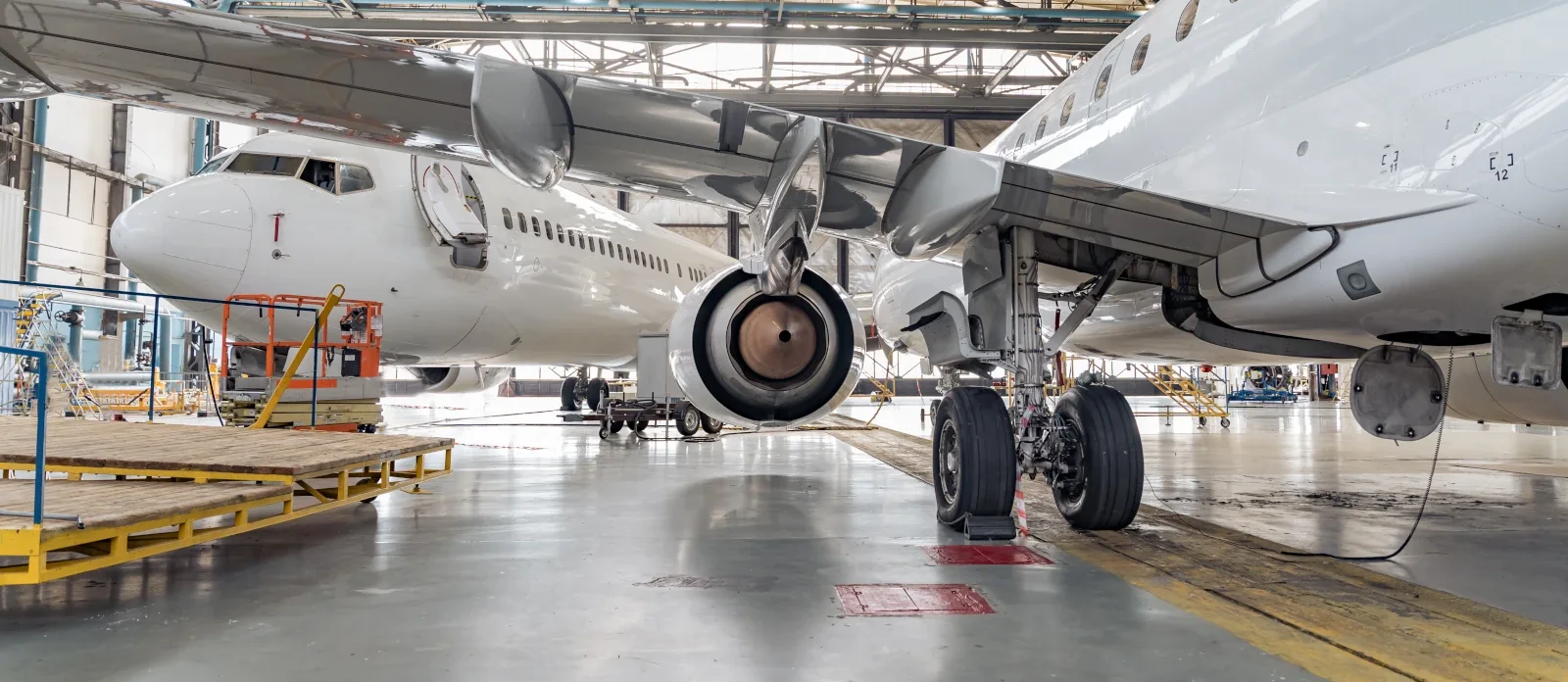 Two passenger jets parked for maintenance in a large hangar, viewed from beneath a wing and landing gear