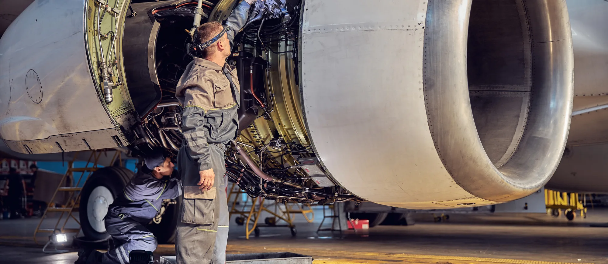 Two aviation mechanics working on an open jet engine in a hangar, one standing and one kneeling by the landing gear